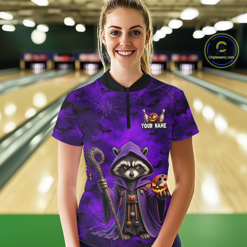 A woman stands in a bowling alley wearing a purple short-sleeve bowling shirt with a cartoon raccoon in a hood holding a pumpkin and staff, plus a black collar and a partial black zipper. The shirt has a customizable text graphic on the chest.