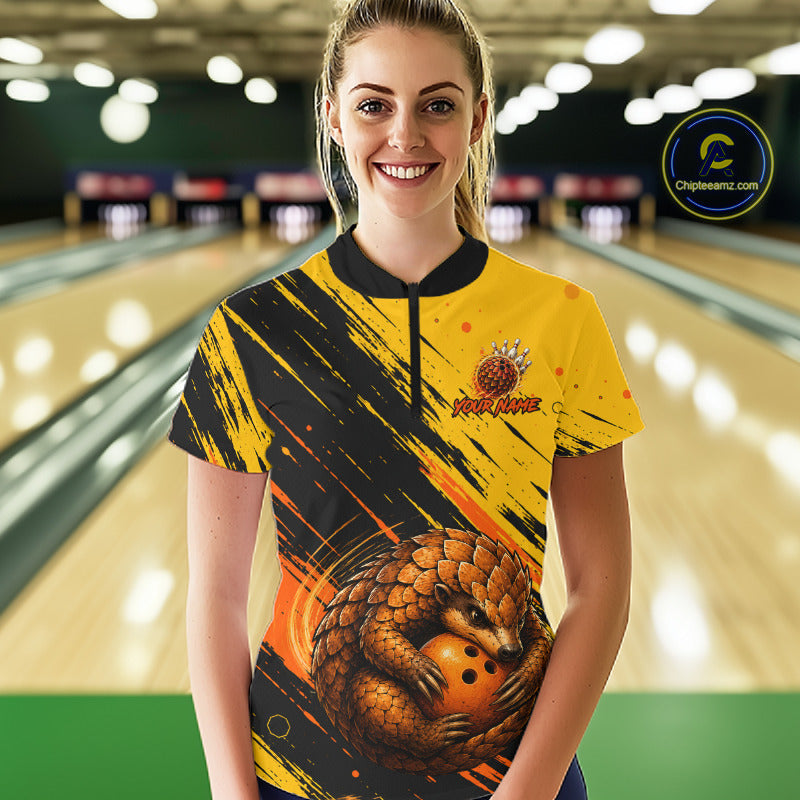 A woman stands in a bowling alley wearing a short-sleeve bowling shirt with a yellow and black paint-splatter design, a large pangolin graphic, and a bowling pin logo on the chest. 
