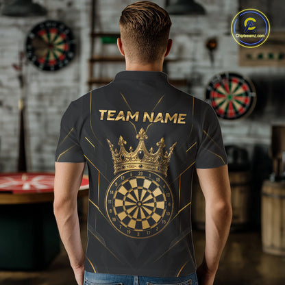 A man viewed from behind wearing a dark gray short-sleeve polo shirt with a large gold crown and dartboard graphic on the back, plus the text 'TEAM NAME'. The shirt is shown in a blurred indoor game room setting with dartboards in the background.