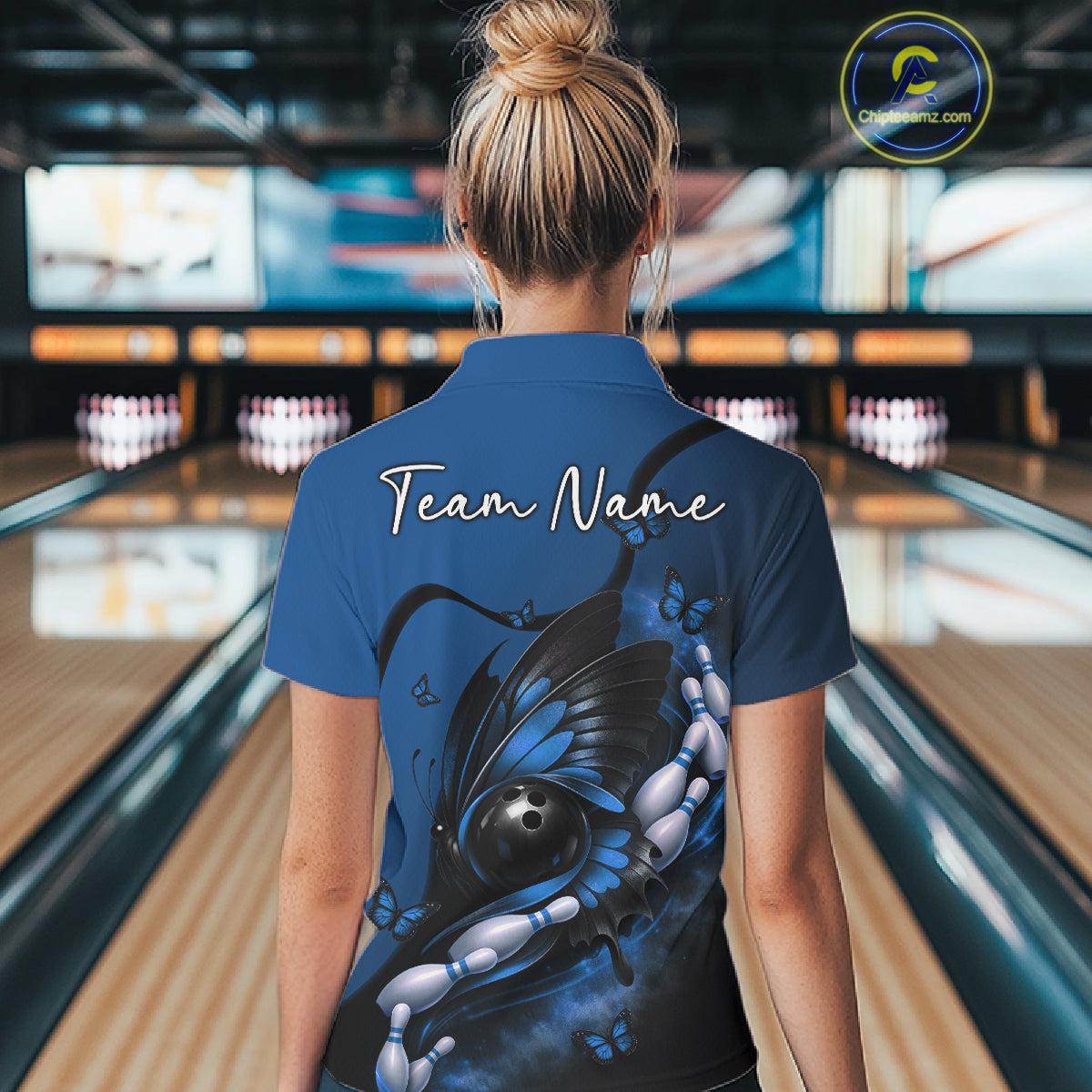 Back view of a female model in a bowling center wearing a blue and black polo shirt featuring a butterfly wrapped around a bowling ball.