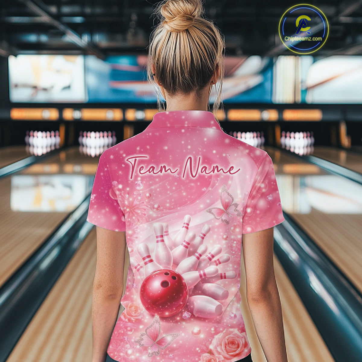 Back view of a female model in a bowling alley wearing a pink quarter zip shirt featuring a bowling ball striking pins with butterflies.