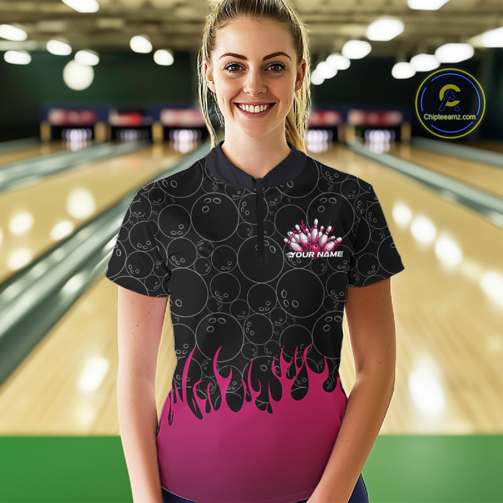 In a bowling lane, front view of a female model wearing a black quarter-zip shirt with vibrant pink flames and bowling ball pattern graphics creating a bold bowling action scene.