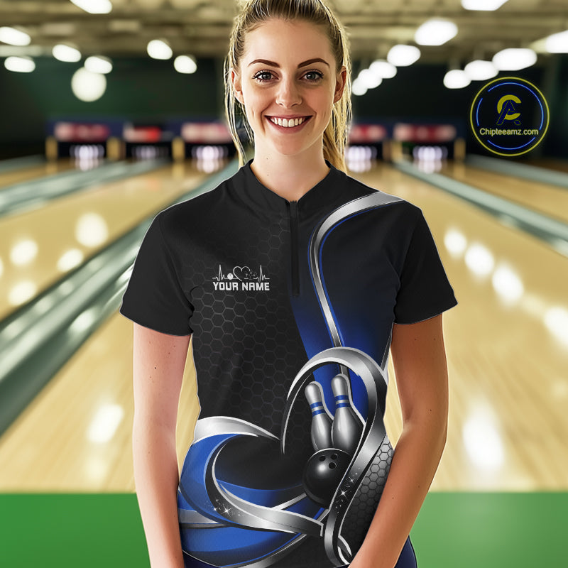 Female model in a bowling alley wearing a black and blue quarter zip shirt, showing a honeycomb pattern and heart motif surrounding a bowling ball and pins.