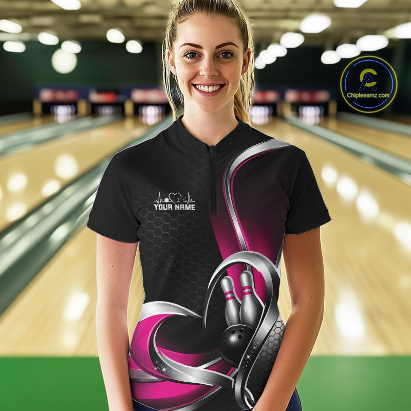 Female model wearing a black and pink bowling quarter zip shirt in a bowling alley, showing a honeycomb pattern with a heart surrounding a black bowling ball and pins.