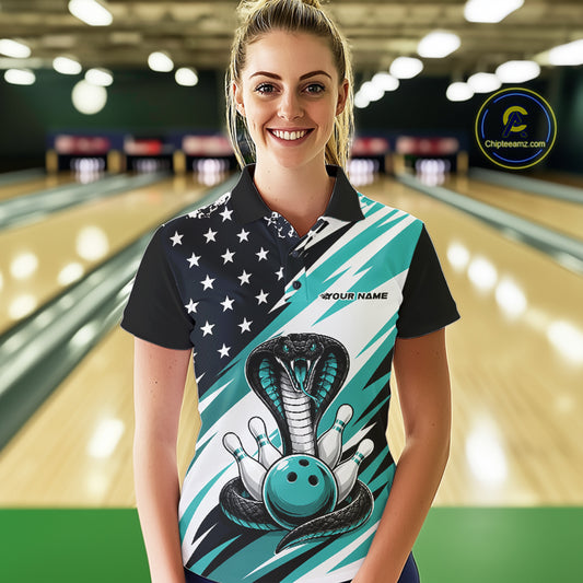 A woman stands in a bowling alley wearing a black, white, and blue bowling shirt with stars, diagonal stripes, a cobra, pins, and an blue ball.