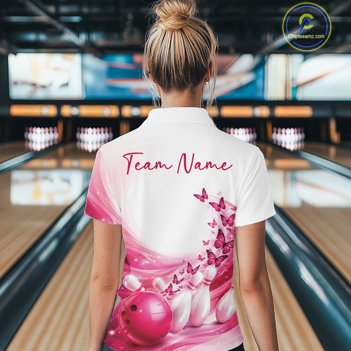Female model wearing a white and pink bowling polo with butterfly artwork and bowling graphics, back view inside a bowling center