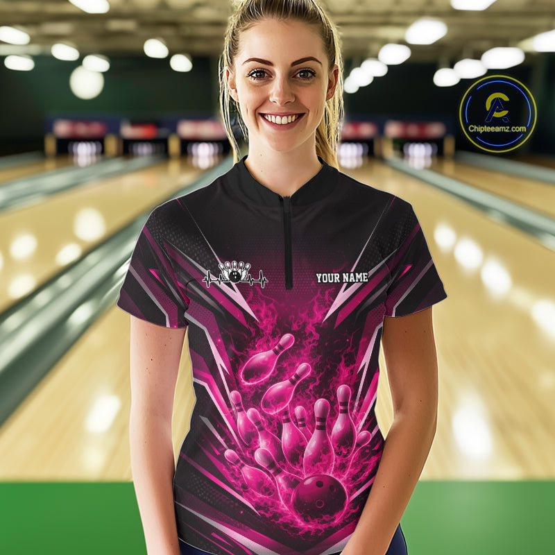 Front view female model at bowling lane wearing black and pink quarter zip shirt showing flaming bowling ball striking pins with energetic fire burst illustration.