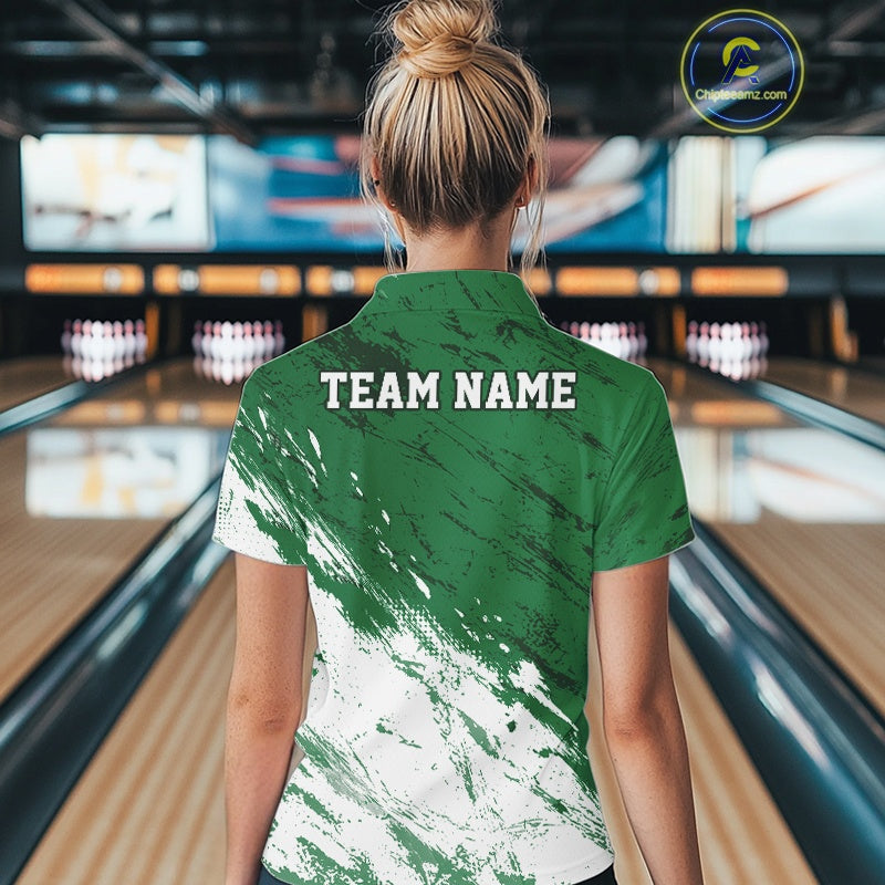 Back view of a female model in a bowling alley wearing a bowling polo shirt featuring a white paint splatter distressed design on a green shirt.
