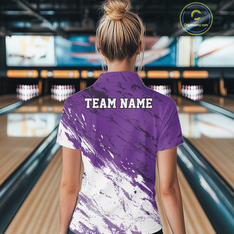 Back view of a female model in a bowling alley wearing a bowling quarter-zip shirt featuring a white paint splatter distressed sportswear design on a purple shirt.
