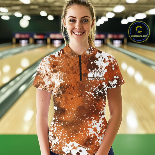 Front view of a female model in a bowling alley wearing a bowling quarter-zip shirt featuring a white paint splatter design on an orange sportswear shirt.

