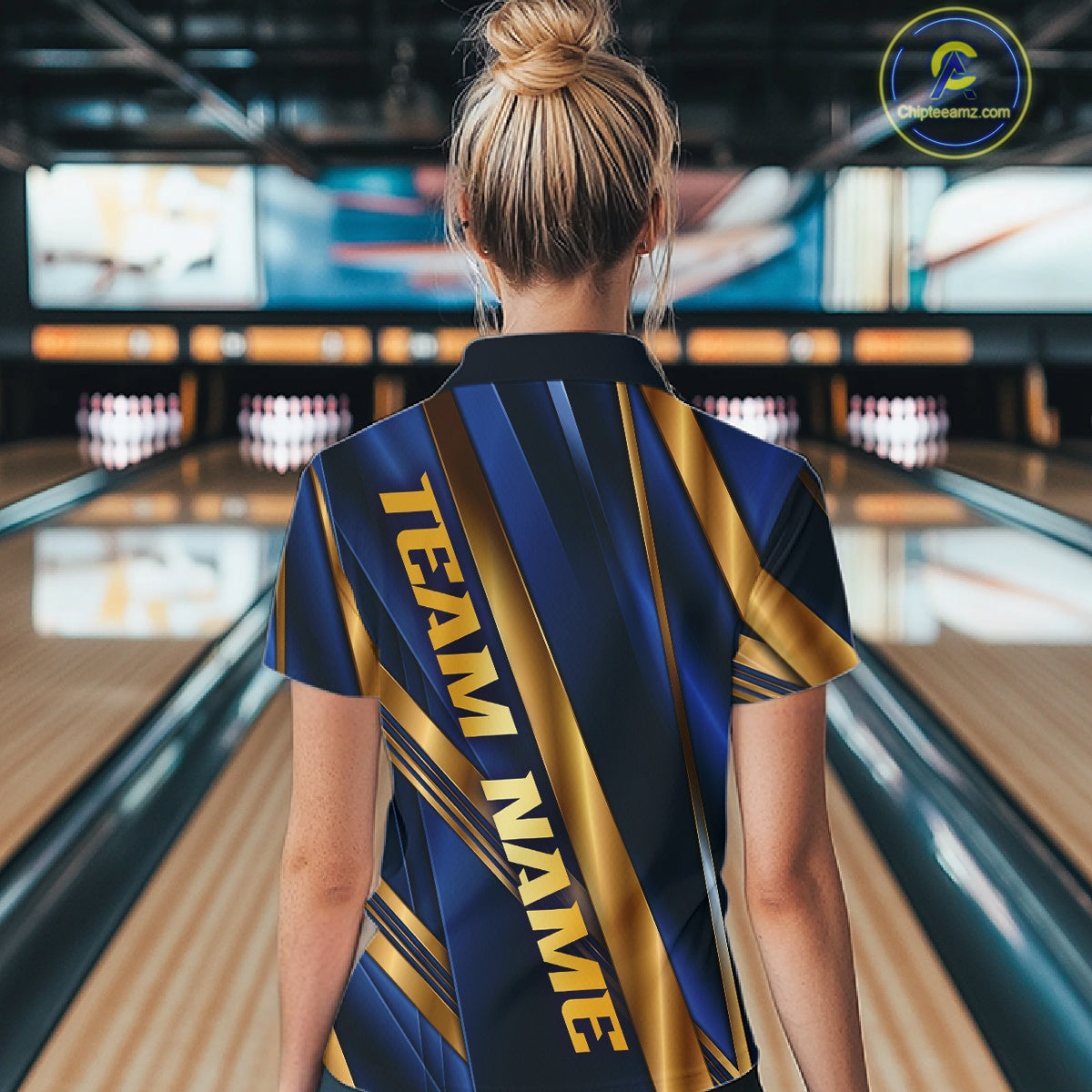A woman viewed from behind wearing a short-sleeve bowling shirt with a navy and gold diagonal stripe pattern and the text 'TEAM NAME' printed vertically on the back. The background shows a blurred bowling alley with lanes and pins.