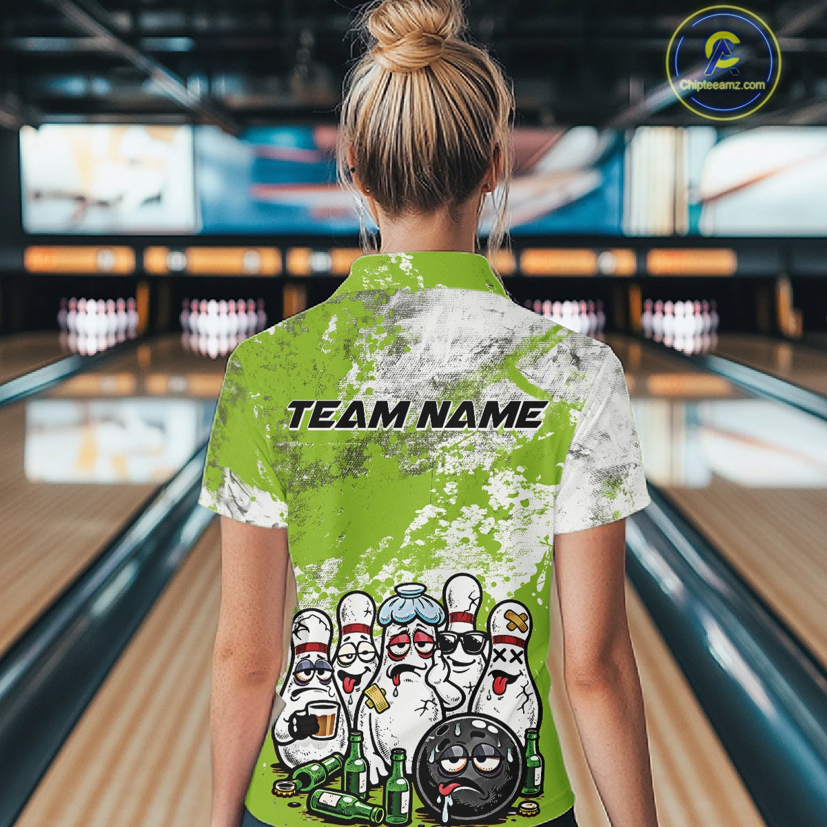 Back view of a female model in a bowling lane wearing a white quarter-zip shirt with green and white grunge splatter pattern and humorous bowling pins illustration.
