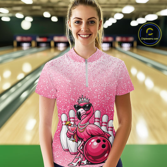 Female model wearing a pink bowling quarter-zip shirt at a bowling lane with a crowned flamingo holding a cocktail and resting against a bowling ball.