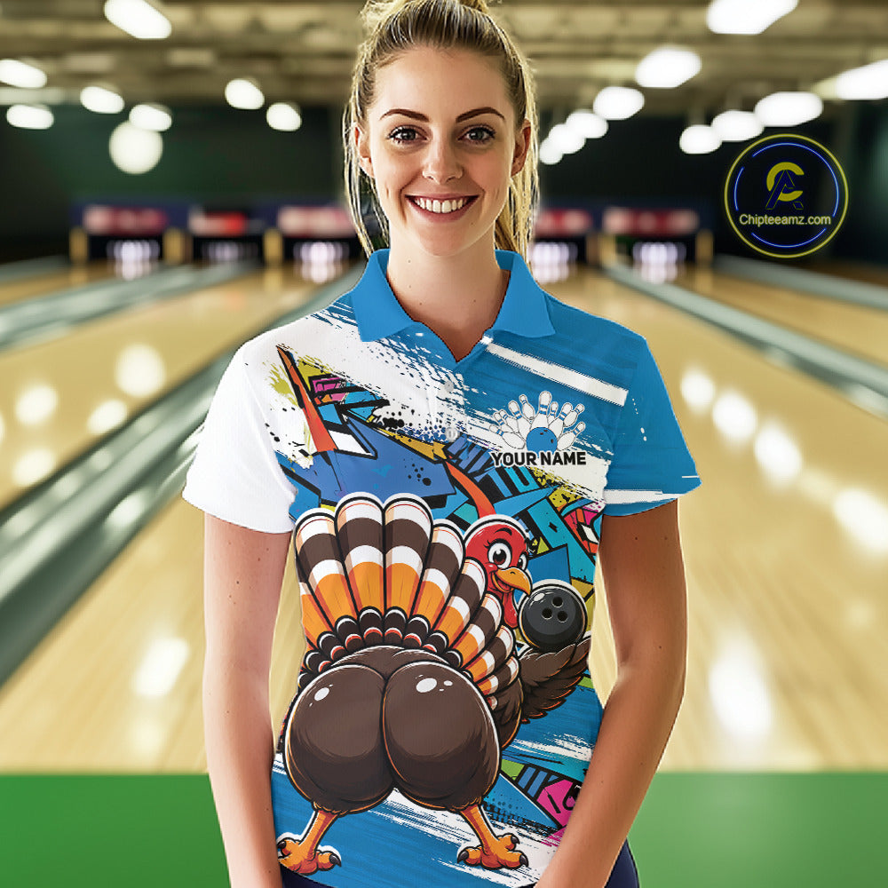 Front view of a female model wearing a blue custom bowling polo shirt with a funny graffiti turkey holding a bowling ball, paint splatter accents, and a personalized name in a bowling alley.
