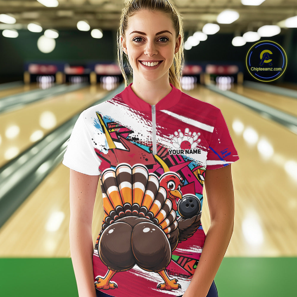 Front view of a happy female model in a bowling alley wearing a red custom quarter zip bowling shirt with a funny graffiti turkey holding a bowling ball, paint splatter accents, and a personalized name.