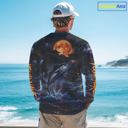 A man stands with his back to the camera wearing a long-sleeve graphic shirt featuring a large jumping fish and a glowing full moon on a dark background, with orange text down the sleeves, set against an ocean backdrop.