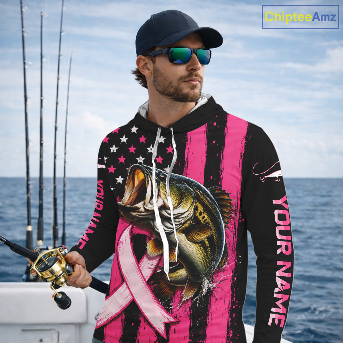 A man wearing a custom pink and black striped hoodie with a bass graphic, a pink ribbon, and 'YOUR NAME' printed on the sleeve, standing on a boat with fishing rods and the ocean in the background