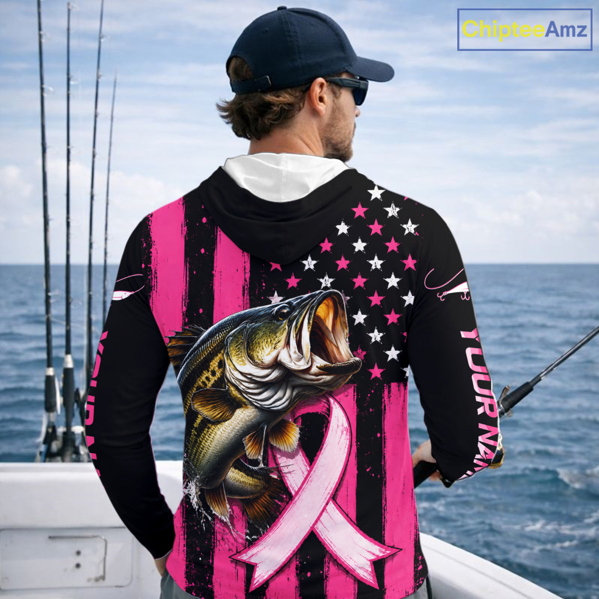 A man on a boat wearing a black and pink long-sleeve hoodie with a large graphic of a bass fish over a pink ribbon and stars-and-stripes design. The photo is taken from behind, showing the back of the hoodie in a real outdoor ocean setting.
