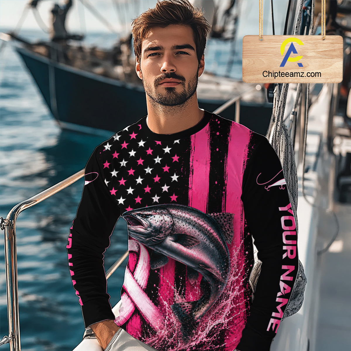 A man wearing a long-sleeve custom crewneck shirt with a pink and black American flag design, a large fish graphic, and 'YOUR NAME' printed on the sleeve, standing on a boat with water and another boat in the background.
