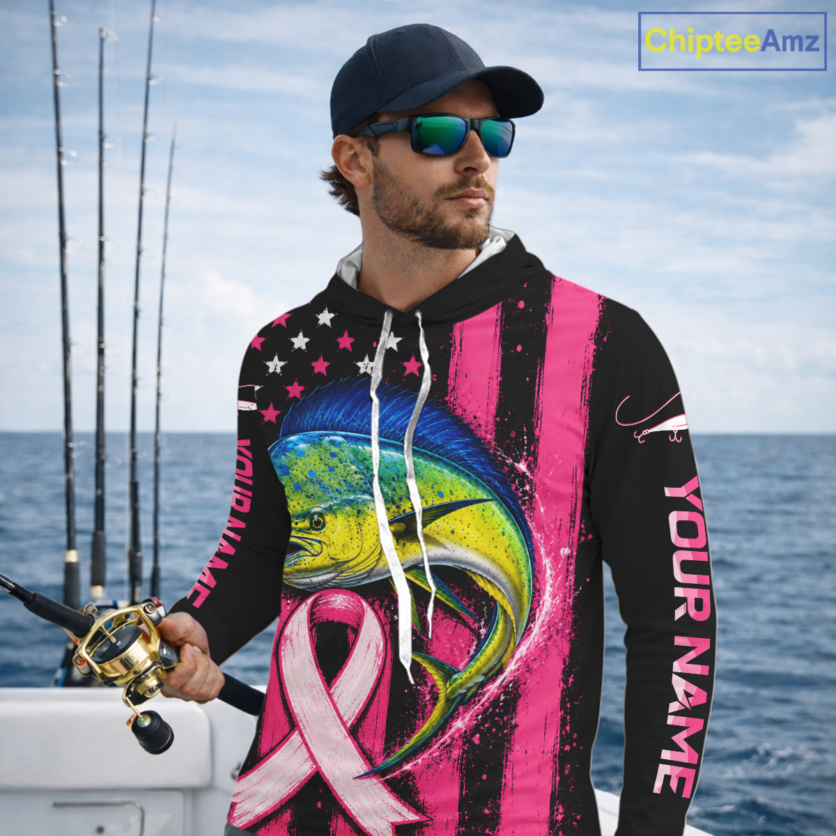 A man wearing a custom black and pink fishing hoodie with a large marlin graphic and pink awareness ribbon, standing on a boat with fishing rods and ocean in the background.
