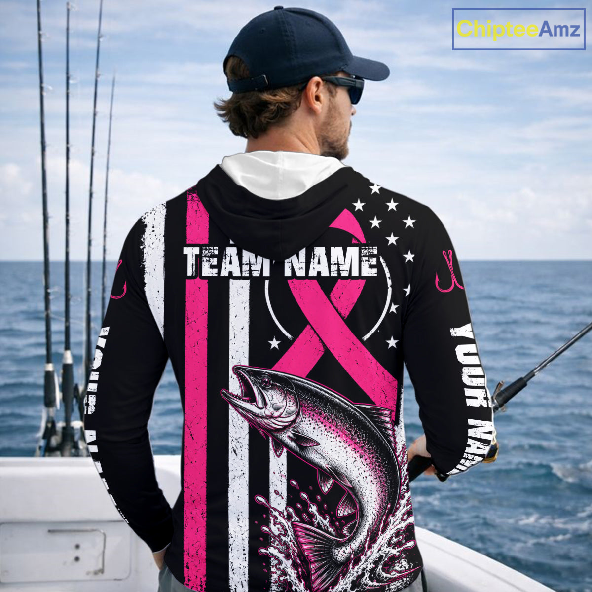 A man on a boat shown from behind wearing a black long-sleeve fishing hoodie with a pink and white striped design, a pink awareness ribbon, a large fish graphic, and customizable text. The background shows the ocean and fishing rods.