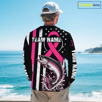 A man stands on a beach facing away from the camera, wearing a black long-sleeve shirt with a large pink and white graphic design featuring a fish, stars, stripes, and customizable text. The background shows the blue ocean and sky.