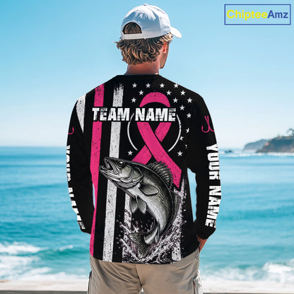 A man standing on a beach viewed from behind, wearing a black long-sleeve shirt with a large pink and white graphic design featuring a fish, stars, stripes, and customizable text. The background shows the blue ocean and sky.