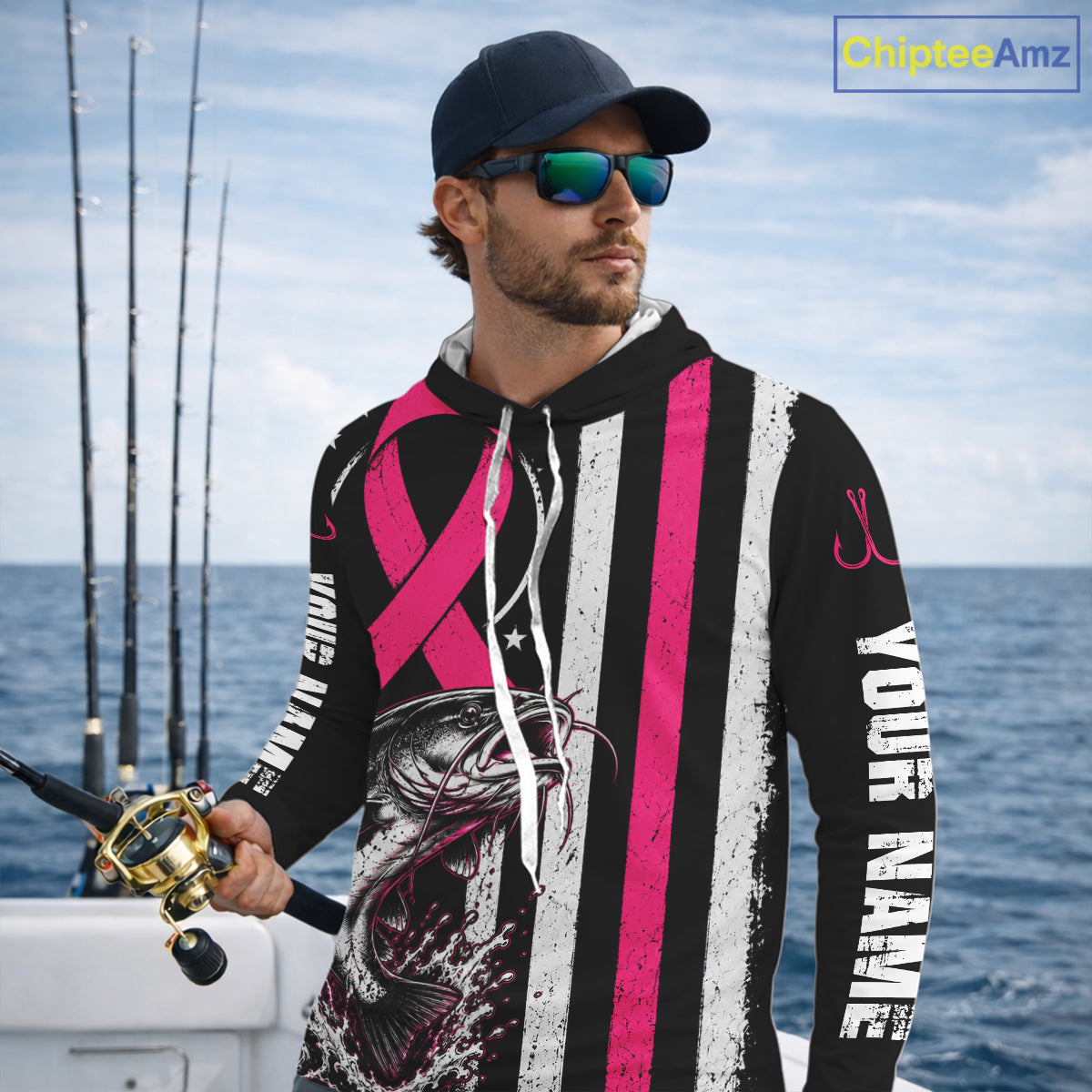 A man on a boat wearing a custom black, white, and pink striped hoodie with a fishing fish graphic and 'YOUR NAME' printed on the sleeve. He is also wearing a navy cap and sunglasses.