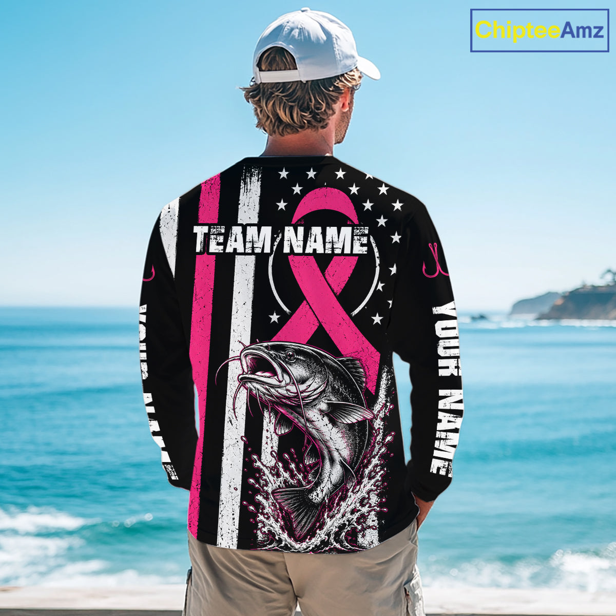 A man stands on a beach with his back to the camera wearing a black long-sleeve shirt with a pink and white patriotic fishing design, including a fish graphic, stars, stripes, and customizable text placeholders.