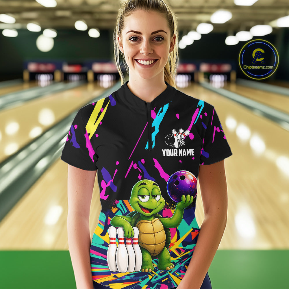 Person wearing a black bowling shirt with colorful design and turtle graphic in a bowling alley.